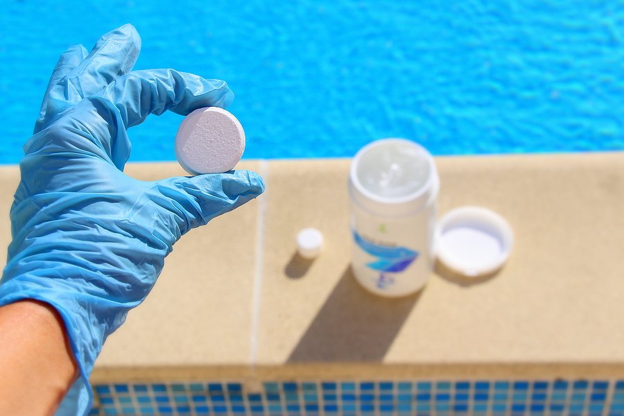Gloved hand holding a pool chlorine tablet near a pool.